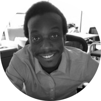 A black and white photo of Gregory McClendon seated in an office chair smiling and wearing an Oxford button-up shirt