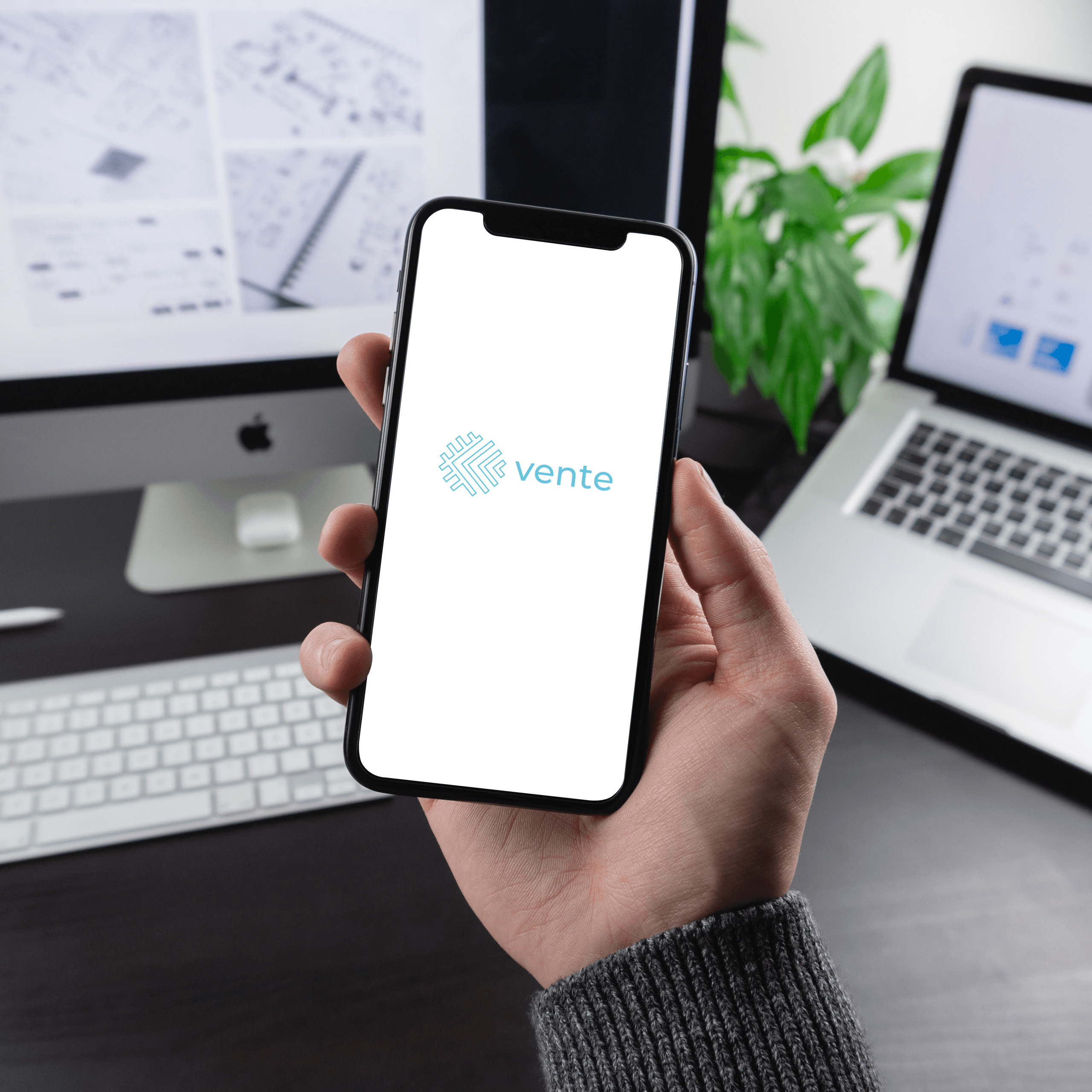 A photo of a man's hand holding an iPhone X with the Vente logo mocked up on it. A desktop computer and laptop are in the background on the desk. 